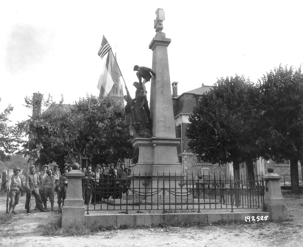 Trois jeunes gens escaladent le Monument aux morts sur la Place de Barenton sous le regard de soldats américains et de civils. Drapeau américain et drapeau français installés sur la statue.