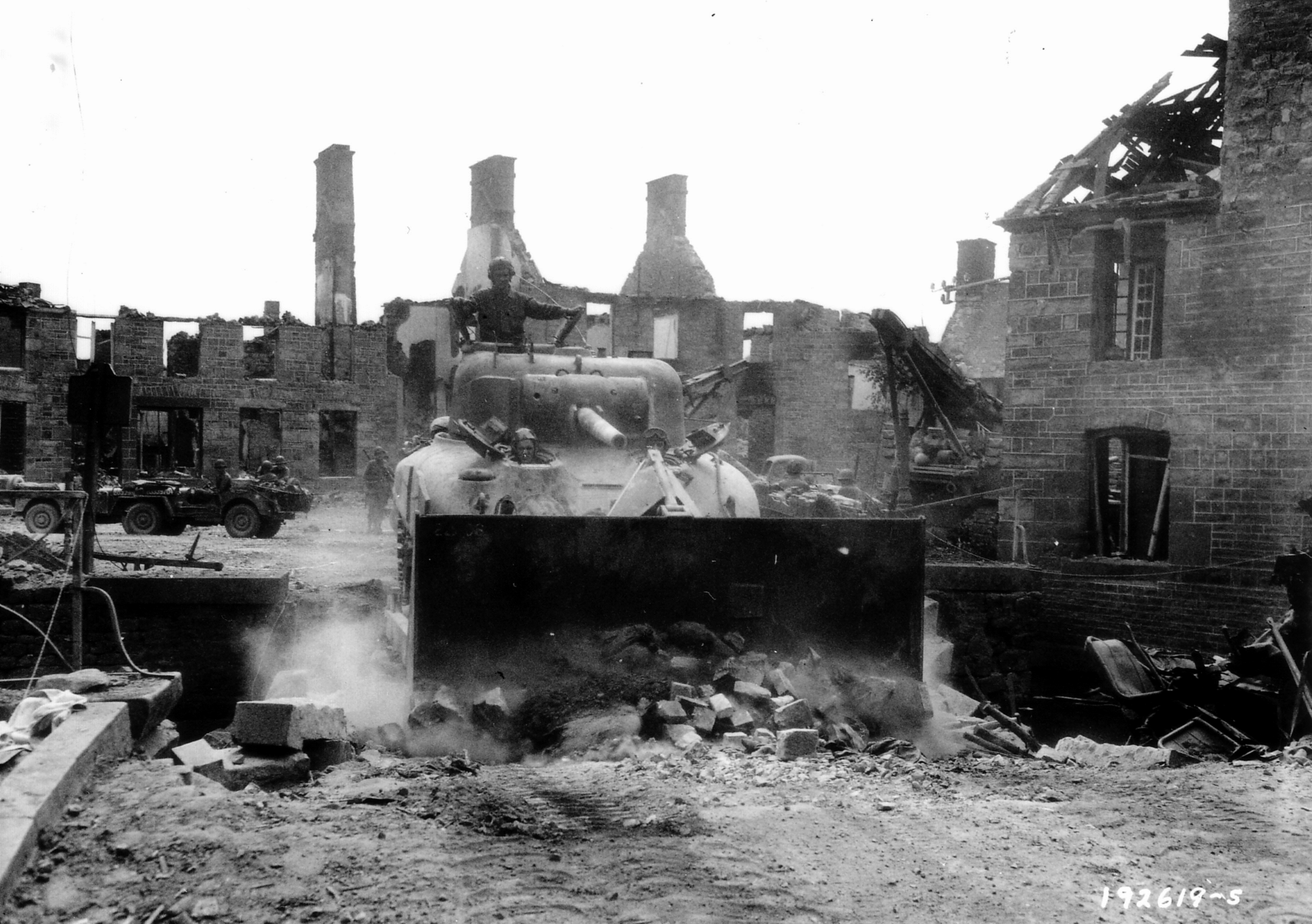 A M4A1 Sherman tank fitted with a bulldozer blade clears rubble from the bridge in the center of the village. The bridge was partially destroyed by an American elements of the 17th Armored Engineer Battalion and 82nd Recon. Bn (2nd US AD) at around 23:00 on the 8th of August during the German Operation Lüttich offensive near Mortain.  This was to prevent units of the 10th SS Panzer advancing the area from using the bridge to further their advance.  On the Right hand side you can just see the rear end of a TRV  The photo credit I have says its the ruins in Lonlay-L'Abbaye on 15 August 1944. This came with the photo. As far as we know this is the correct caption date time and place Source Facebook page 2nd Amorored Maintanance Battalion