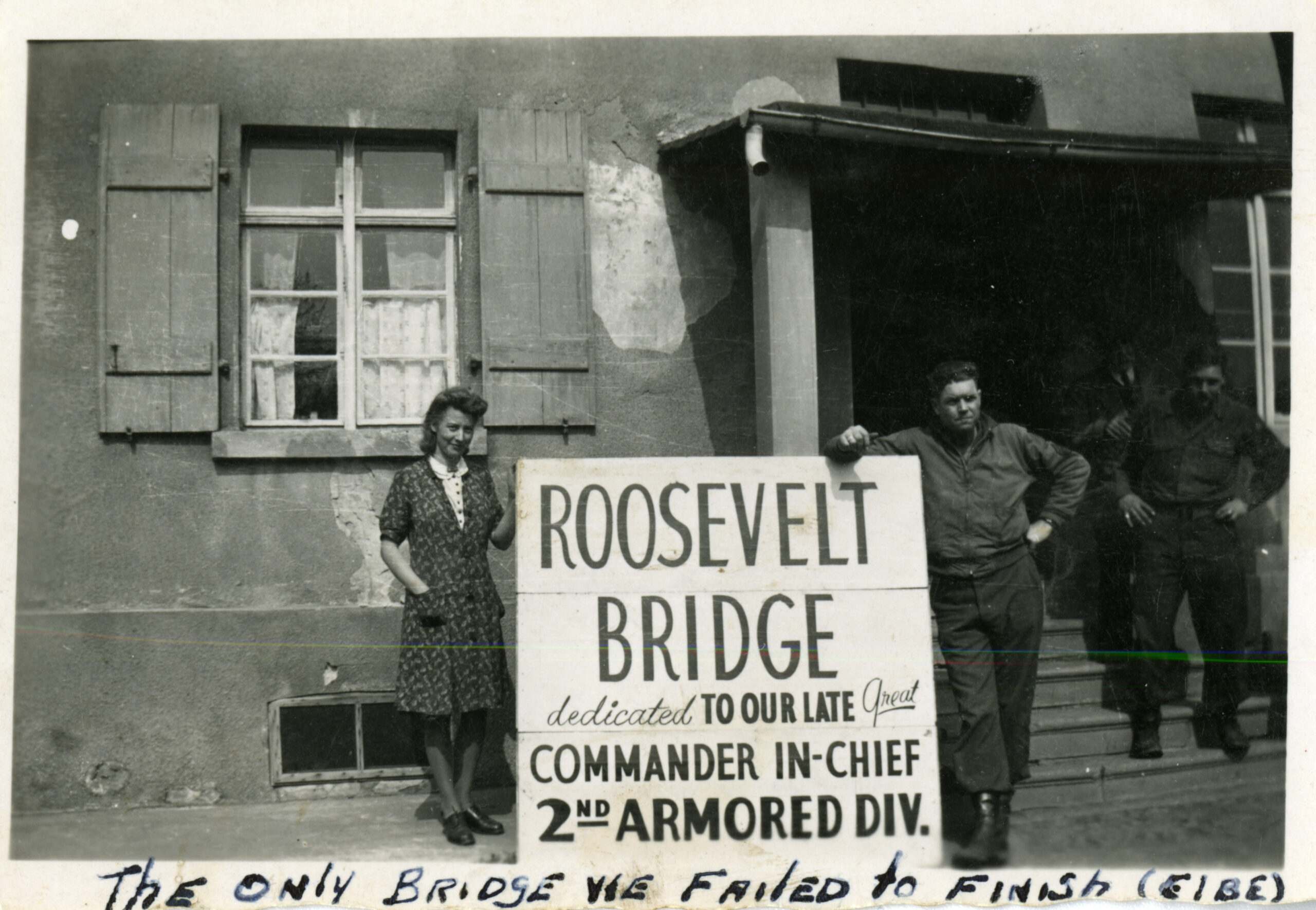 American serviceman and a German woman posing with a sign that reads "Roosevelt Bridge dedicated to our late great Commander in-chief 2nd Armored Div." ; another serviceman is standing on the steps of the building in the background. Personal caption on front: "The only bridge we failed to finish (Elbe) ." Westerhusen, Germany. April 1945